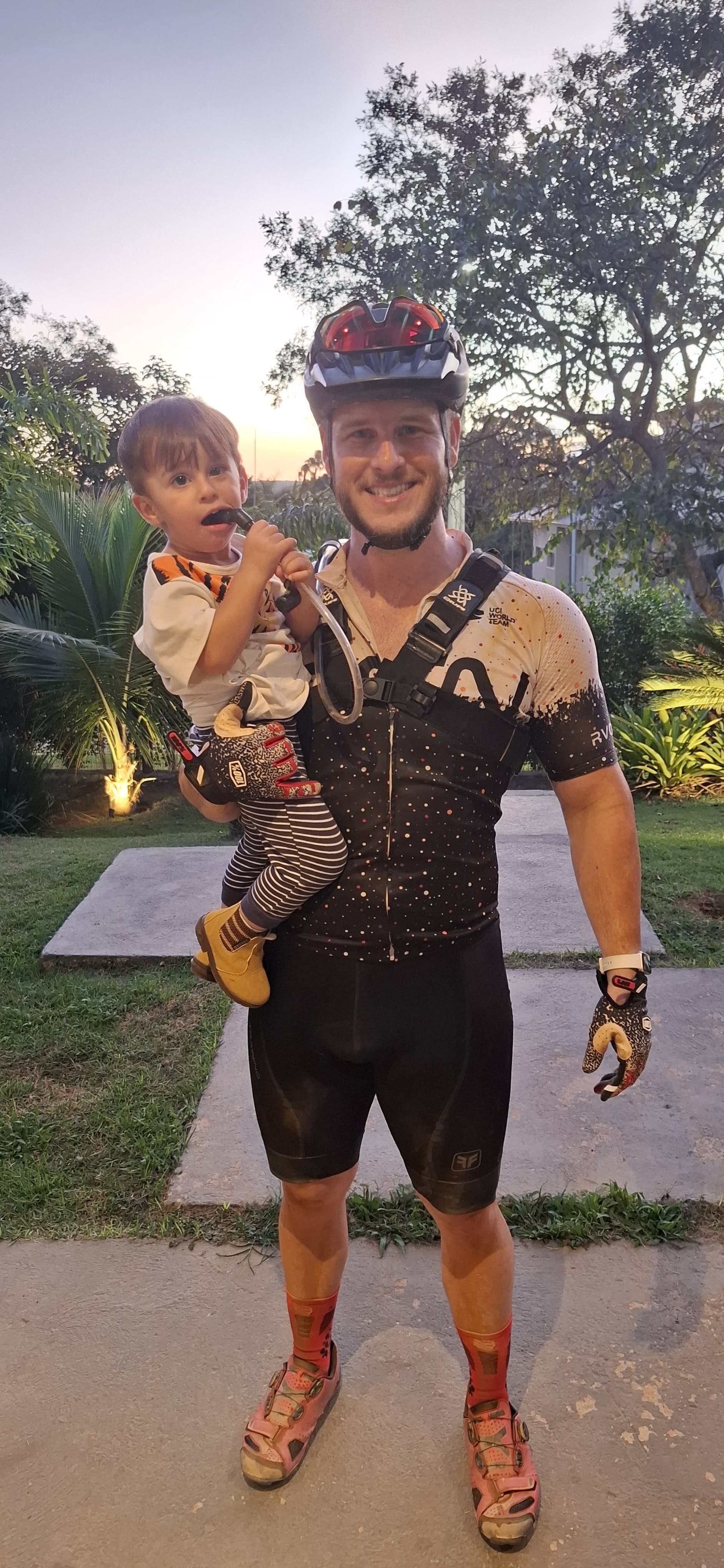 Felipe Malafaia com seu filho no colo após uma pedalada, usando capacete e roupa de ciclismo